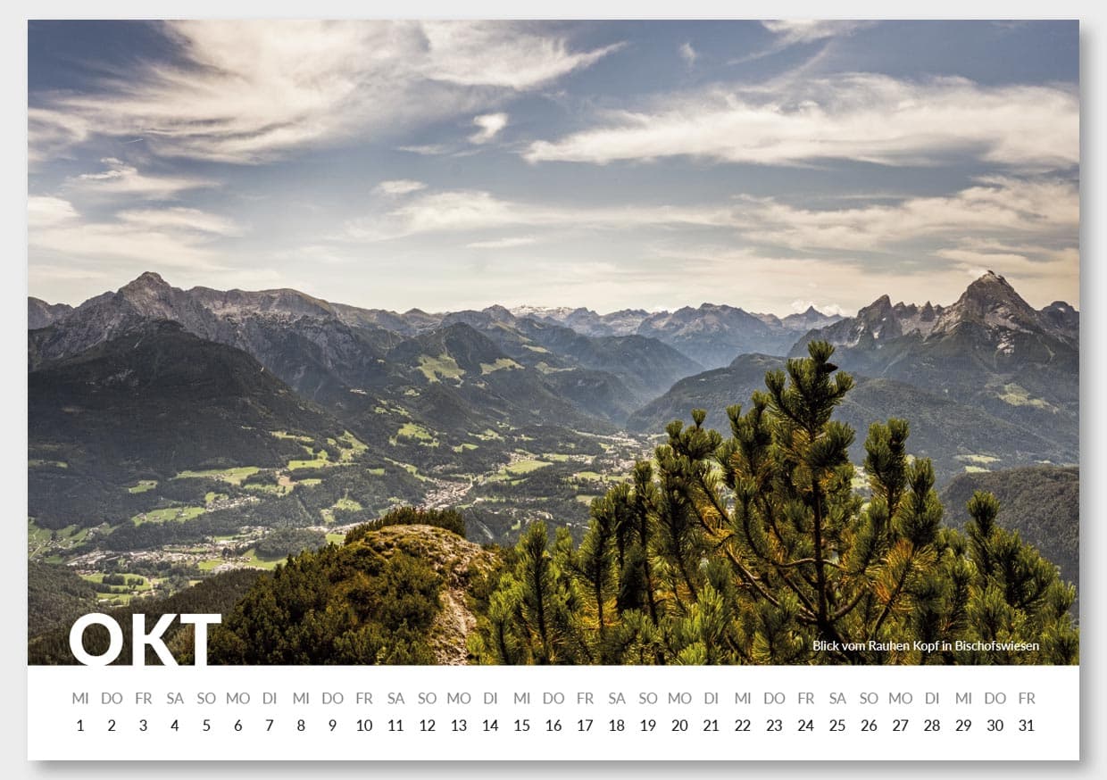 Blick vom Rauhen Kopf in Bischofswiesen auf eine Berglandschaft mit bewaldeten Hügeln und mächtigen Bergen im Hintergrund, unter einem bewölkten Himmel.