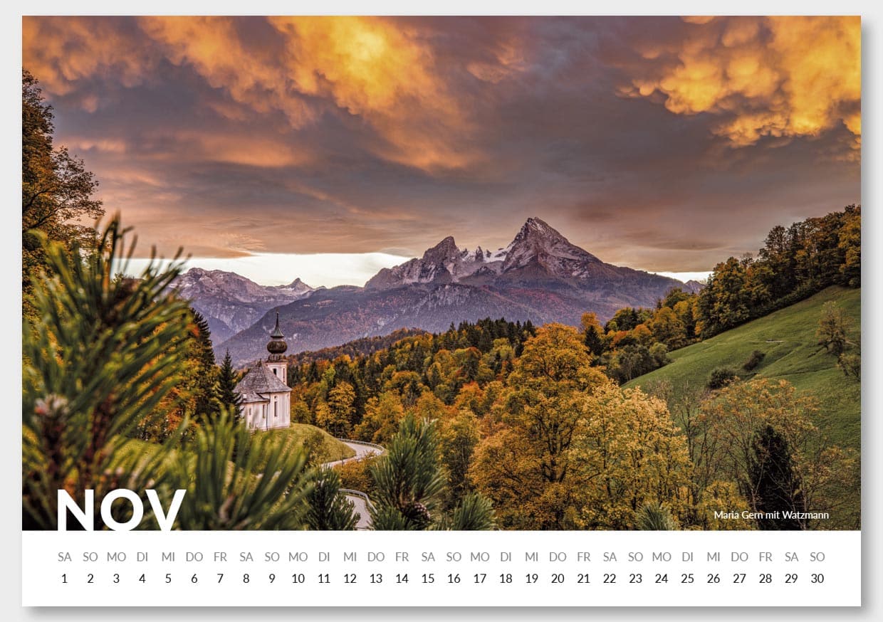 Ein Landschaftsbild mit einer kleinen Kirche im Vordergrund, umgeben von bunten Herbstbäumen, mit hohen Bergen im Hintergrund und einem dramatischen, orange-gelben Himmel. Im unteren rechten Bereich steht der Text "Maria Gern mit Watzmann".