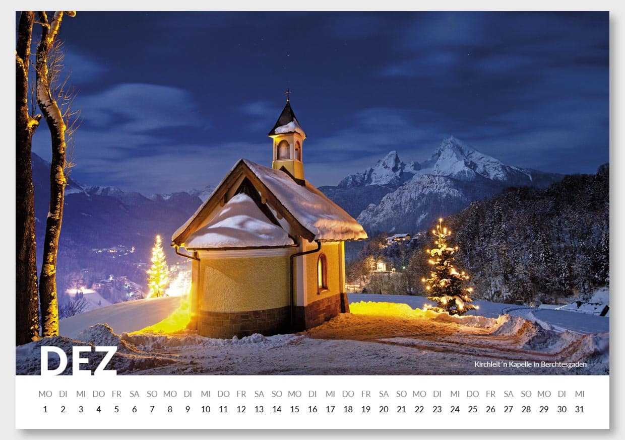 Ein kleiner, weihnachtlich beleuchteter Kirchturm in einer verschneiten Winterlandschaft vor einer Reihe von Bergen bei Nacht, mit einem bewachsenen, beleuchteten Baum im Vordergrund.