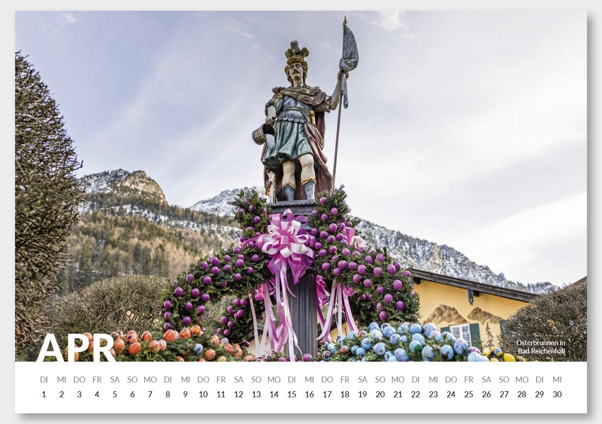 Eine Osterfigur auf einem Weihnachtskranz vor hoher, bewaldeter Bergkette im Hintergrund, mit einem Haus im rechten Hintergrund, dekoriert mit pinkfarbenen und blauen Ostereiern, für den Monat April in einem Kalender.